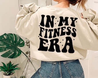 May include: A cream-colored sweatshirt with the text "IN MY FITNESS ERA" in black retro-style lettering with stars on a woman's back.