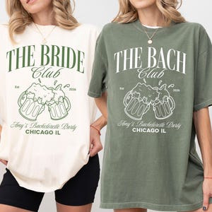 Könnte beinhalten: Zwei T-Shirts, eines in Elfenbein und eines in Moosgrün, mit passenden Grafiken. Das elfenbeinfarbene Shirt trägt die Aufschrift "THE BRIDE Club" mit Bierkrügen und "Amy's Bachelorette Party CHICAGO IL". Das moosgrüne Shirt trägt die Aufschrift "THE BACH Club" mit ähnlichen Grafiken.