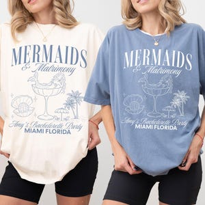 以下が含まれることがあります： アイボリーとジーンズブルーの半袖Tシャツ2枚。各シャツには「MERMAIDS & Matrimony」、「Amy's Bachelorette Party」、「MIAMI FLORIDA」というテキストと、グラスに入った人魚、貝殻に入った真珠、ヤシの木のイラストが描かれたグラフィックプリントが施されています。