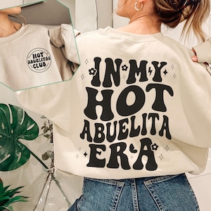 May include: Sand-colored sweatshirt with the text "IN MY HOT ABUELITA ERA" in black, retro-style lettering. The front of the sweatshirt has a small circular logo that says "HOT ABUELITAS CLUB".