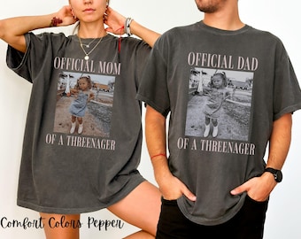 May include: Two charcoal gray short-sleeve t-shirts featuring a central rectangular graphic with a black and white photograph of a child wearing overalls and boots. The text reads OFFICIAL MOM OF A THREENAGER and OFFICIAL DAD OF A THREENAGER.
