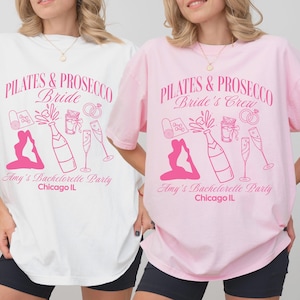 May include: Two white t-shirts with pink text and graphics. The text on the left shirt says "Pilates & Prosecco Bride" and the text on the right shirt says "Pilates & Prosecco Bride's Crew". Both shirts have a graphic of a person doing pilates with a bottle of prosecco.