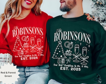 Può includere: Felpe rosse e verde foresta con la scritta "The Robinsons" e "Our First Christmas & Mr. & Mrs. EST. 2025". Il design include un albero di Natale, calici di champagne e fedi nuziali.
