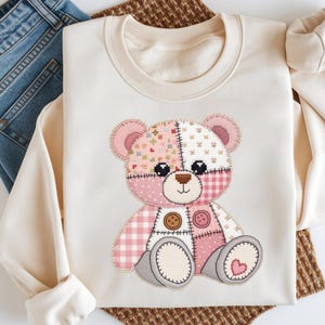 Könnte beinhalten: Cremefarbenes Sweatshirt mit einem Patchwork-Teddybären-Design. Der Bär hat rosa, weiße und gemusterte Flicken, Knopfakzente und ein Herzdetail. Das Sweatshirt ist gefaltet, mit blauen Jeans sichtbar.