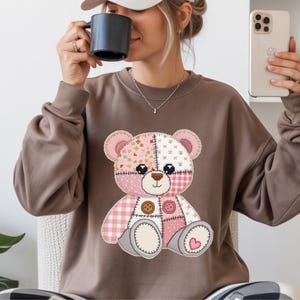 May include: A brown sweatshirt with a patchwork teddy bear design. The bear has pink, white, and patterned patches, with button details and a heart. The sweatshirt is a neutral color, suitable for casual wear.