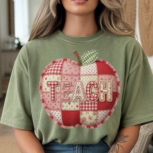 Op de afbeelding: Saliegroen t-shirt met een groot appelontwerp. De appel is gemaakt van patchworkstof in rode, roze en witte patronen, met het woord "TEACH" in het midden. De appel heeft een bruine steel en een groen blad.