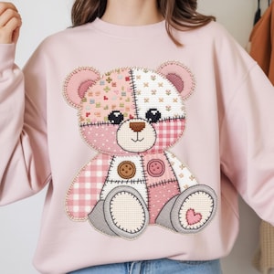 May include: A pale pink sweatshirt with a patchwork teddy bear design. The bear has a brown nose, black eyes, and is made of various pink and white patterned fabrics, including gingham and floral prints. Two brown buttons and a heart detail the design.