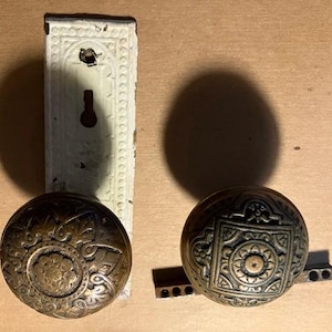 May include: Two antique brass door knobs with intricate floral and geometric designs. The knobs are attached to a white porcelain backplate.