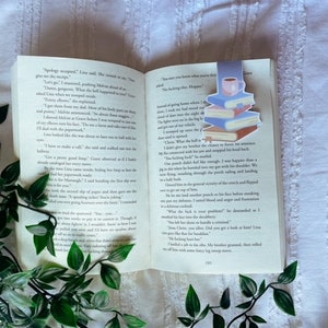 May include: An open book with text, featuring a colourful bookmark depicting a stack of books and a coffee cup. Green leafy vines are draped around the book. The book is on a white, textured surface.