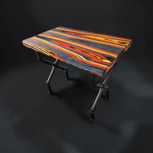 May include: A rectangular table featuring a tabletop made of multi-colored layered wood with vibrant red, yellow, blue, and black stripes. The table is supported by a base constructed from dark grey industrial metal pipes and fittings.
