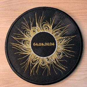 May include: Black circular patch with gold and white embroidery of a sun with rays. The date "04.06.2024" is embroidered in white inside the sun.