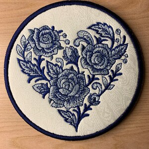 May include: A white circular patch with a navy blue embroidered design of three roses in a heart shape.