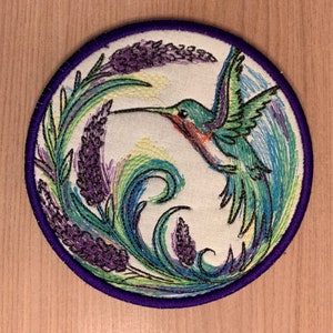 May include: A round embroidered patch with a purple border featuring a green hummingbird with a long beak flying around a swirl of green and blue leaves and purple flowers.