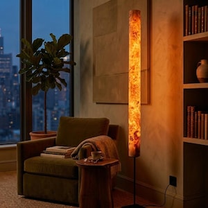 May include: A tall, cylindrical floor lamp featuring a warm, glowing light that illuminates a complex, swirling wood grain pattern. The lamp is supported by a thin, black metal pole and a square base, casting a soft amber light across the room.
