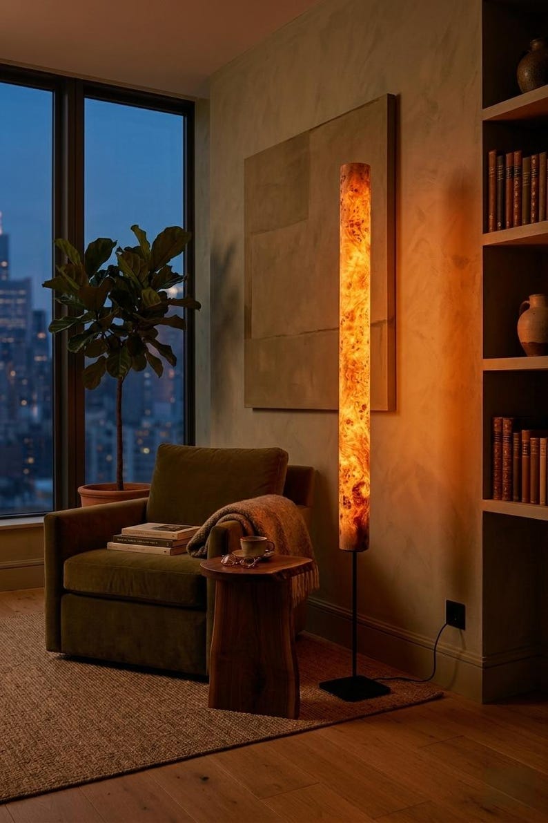 May include: A tall, cylindrical floor lamp featuring a warm, glowing light that illuminates a complex, swirling wood grain pattern. The lamp is supported by a thin, black metal pole and a square base, casting a soft amber light across the room.