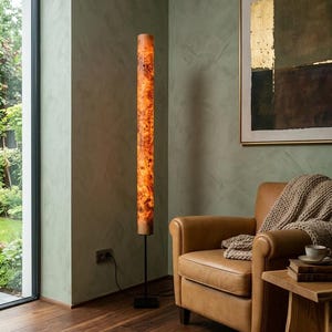 May include: A tall, cylindrical floor lamp featuring a warm, illuminated burl wood veneer shade with a distinct natural grain pattern. The lamp is supported by a slender black metal pole and a square black base, standing upright on a wooden floor.