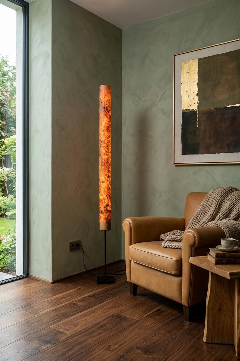 May include: A tall, cylindrical floor lamp featuring a warm, illuminated burl wood veneer shade with a distinct natural grain pattern. The lamp is supported by a slender black metal pole and a square black base, standing upright on a wooden floor.