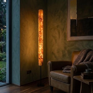 May include: A tall, cylindrical floor lamp featuring a warm, illuminated amber-toned wood grain pattern. The lamp is supported by a slender black metal pole and a square black base, casting a soft, golden glow in a dimly lit room.
