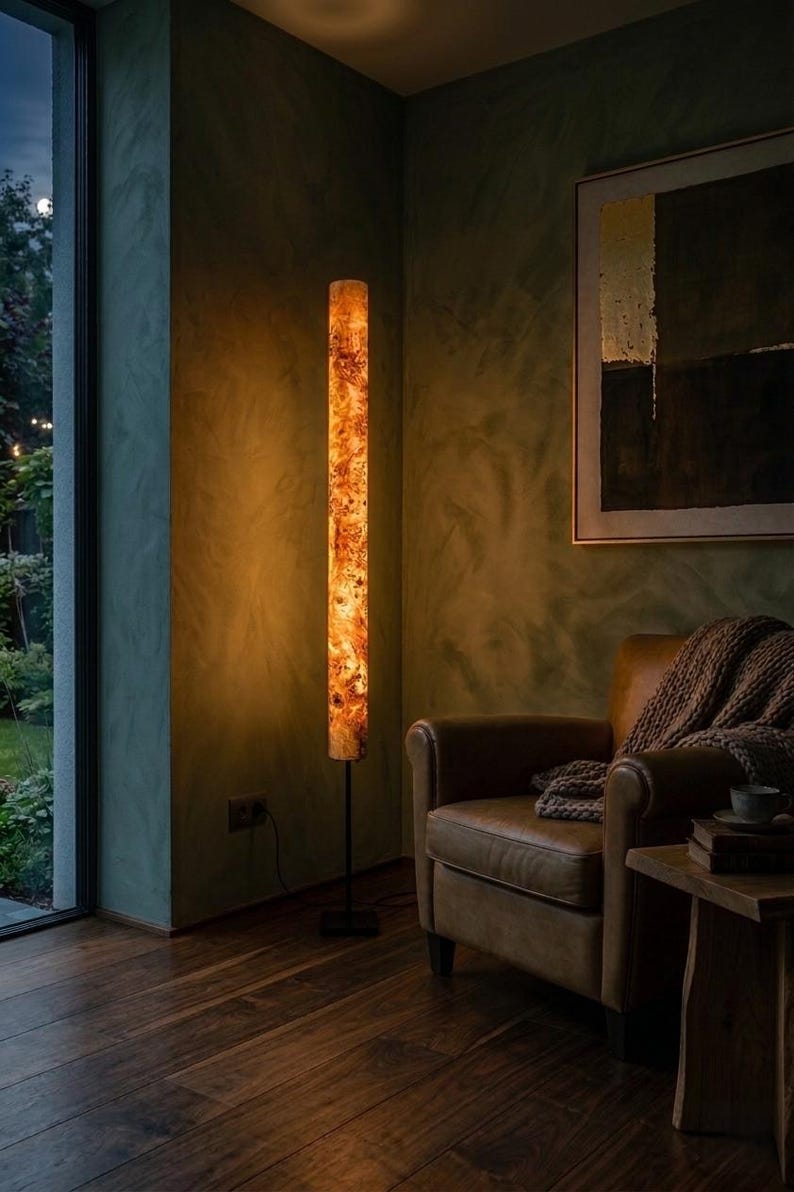 May include: A tall, cylindrical floor lamp featuring a warm, illuminated amber-toned wood grain pattern. The lamp is supported by a slender black metal pole and a square black base, casting a soft, golden glow in a dimly lit room.