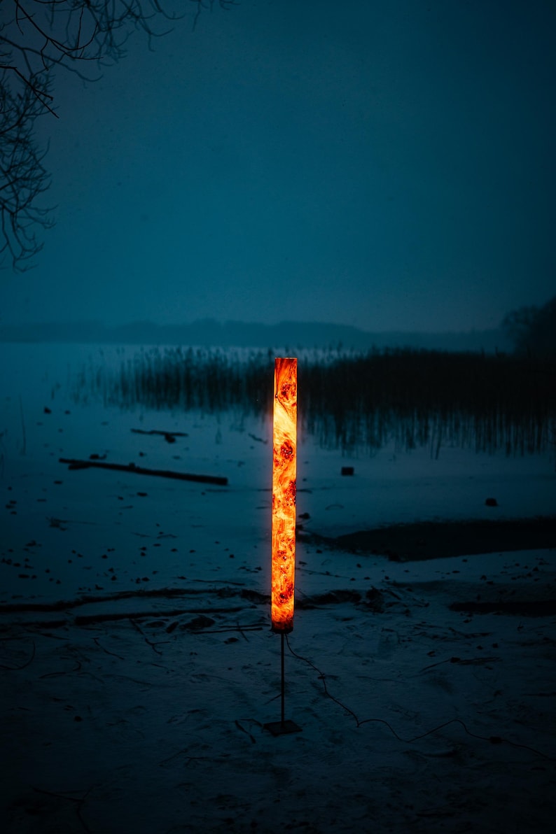 May include: A tall, cylindrical floor lamp featuring a warm, glowing orange and amber wood grain pattern. The lamp is mounted on a thin, dark metal rod attached to a square base, emitting a bright, vertical column of light in a dark environment.