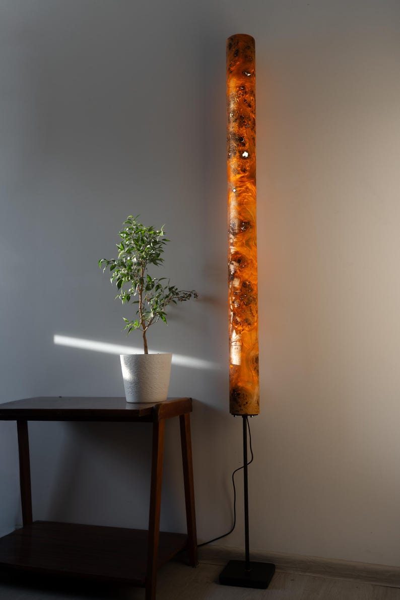 May include: A tall, cylindrical floor lamp featuring a glowing, translucent amber resin body embedded with dark, textured wood burl sections. The lamp is supported by a thin black metal pole and a square base, emitting a warm, diffused light.
