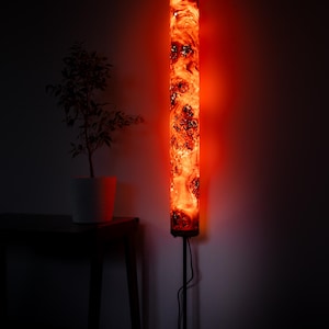 May include: A tall, cylindrical floor lamp featuring a translucent resin body with swirling orange and red patterns that glow intensely. The lamp is mounted on a thin, dark metal stand and includes a black power cord extending from the base.