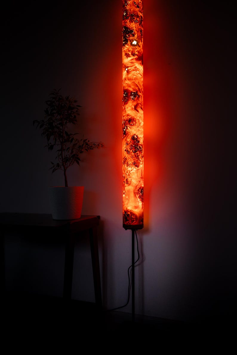 May include: A tall, cylindrical floor lamp featuring a translucent resin body with swirling orange and red patterns that glow intensely. The lamp is mounted on a thin, dark metal stand and includes a black power cord extending from the base.