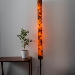May include: A tall, cylindrical floor lamp featuring a glowing orange resin body with dark, intricate wood burl inclusions. The lamp is supported by a thin black metal pole and a square black base, emitting a warm, ambient light from within.