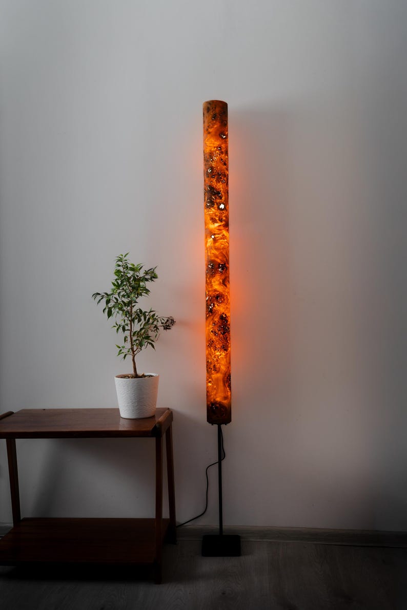 May include: A tall, cylindrical floor lamp featuring a glowing orange resin body with dark, intricate wood burl inclusions. The lamp is supported by a thin black metal pole and a square black base, emitting a warm, ambient light from within.