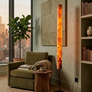 May include: A tall, cylindrical floor lamp featuring a warm, glowing light source encased in a textured, amber-toned wood burl veneer. The lamp is supported by a slender black metal pole and a square black base, standing upright on a floor.