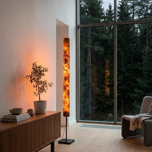 May include: A tall, cylindrical floor lamp with a glowing, marbled orange and black resin shade. The lamp features a slender black metal pole and a square black base, emitting a warm, ambient light that illuminates the surrounding space.