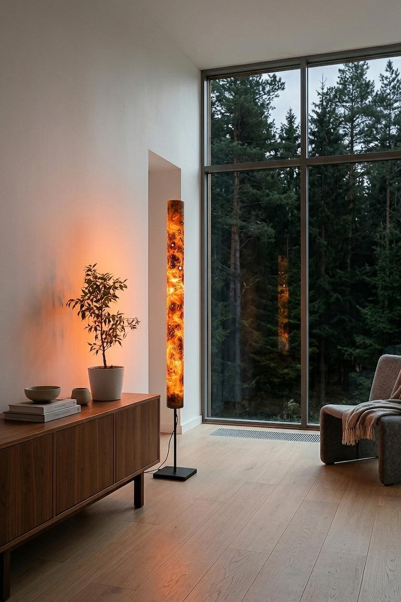May include: A tall, cylindrical floor lamp with a glowing, marbled orange and black resin shade. The lamp features a slender black metal pole and a square black base, emitting a warm, ambient light that illuminates the surrounding space.