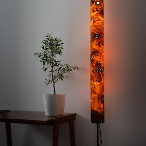 May include: A tall, cylindrical floor lamp featuring a glowing, amber-colored resin and wood burl body with intricate, swirling patterns. The lamp is mounted on a thin, black metal stand and emits a warm, diffused light from within its structure.