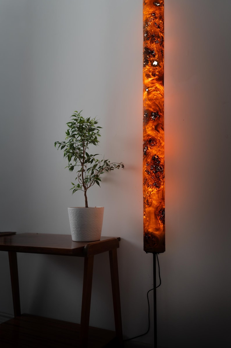 May include: A tall, cylindrical floor lamp featuring a glowing, amber-colored resin and wood burl body with intricate, swirling patterns. The lamp is mounted on a thin, black metal stand and emits a warm, diffused light from within its structure.