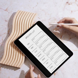 Może przedstawiać: Czarny tablet z białym ekranem, na którym wyświetlony jest szablon listy zakupów. Lista jest uporządkowana według dni tygodnia, z miejscem na wpisanie składników. Biały rysik leży na stole obok tabletu.