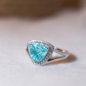 May include: A silver ring with a triangular, light blue gemstone, encircled by small, clear stones. The ring's band has a split design. The gemstone has a textured appearance.