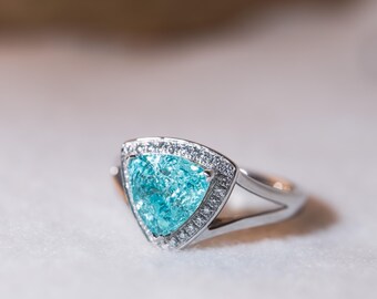 May include: A silver ring featuring a triangular, light blue gemstone, surrounded by a halo of small, clear stones. The ring's band has a split design. The gemstone has a textured appearance.