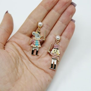 May include: Dangle earrings featuring a mouse and a nutcracker design. The mouse earring has a grey body, pink jacket, and a pearl necklace. The nutcracker earring has a pink and white uniform. Both earrings have a pearl stud.