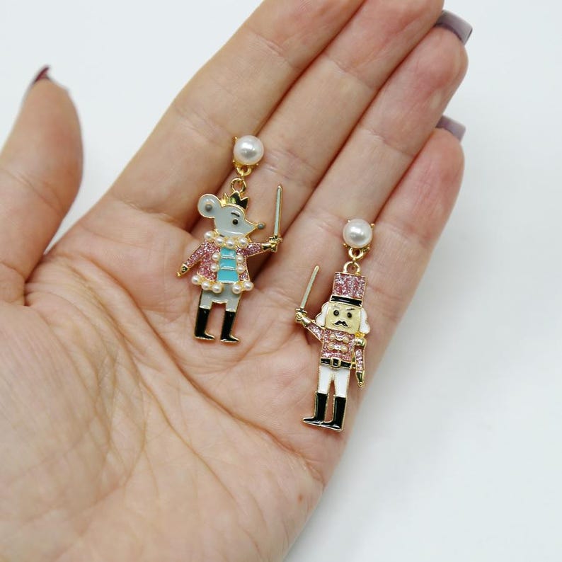 May include: Dangle earrings featuring a mouse and a nutcracker design. The mouse earring has a grey body, pink jacket, and a pearl necklace. The nutcracker earring has a pink and white uniform. Both earrings have a pearl stud.