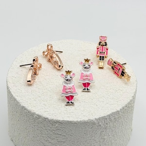 May include: A set of six stud earrings featuring holiday-themed designs including two ballet slippers in rose gold, two nutcracker figures in pink and gold, and two mouse characters wearing pink coats and gold crowns with white and pink enamel details.