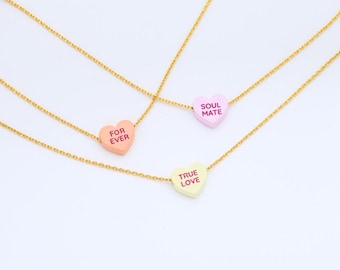 Op de afbeelding: Drie goudkleurige kettingkettingen, elk met een hartvormige hanger. De hangers zijn pastelkleuren: perzik, lavendel en lichtgroen. Elk hart heeft een andere tekst: "FOR EVER", "SOUL MATE" en "TRUE LOVE".