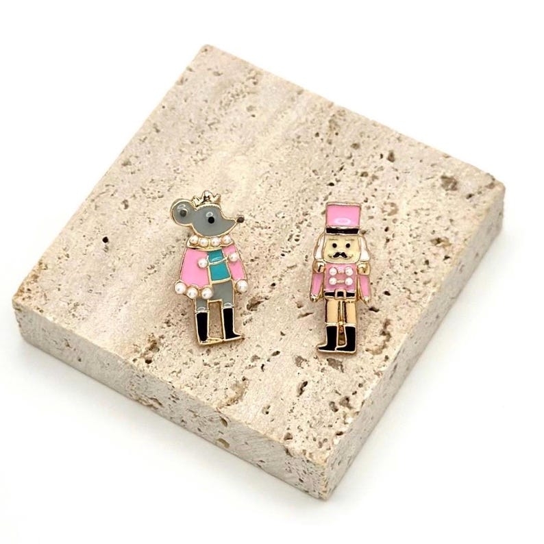 May include: Two enamel pins: a gray mouse wearing a pink coat and a gold crown, and a pink and gold nutcracker soldier. Both pins have black accents and are set on a textured, light-colored surface.