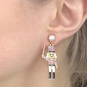 May include: Nutcracker earrings with a pearl stud top. The earrings feature a gold-coloured nutcracker soldier with pink and white accents, holding a sword. The nutcracker has a black moustache and black boots.