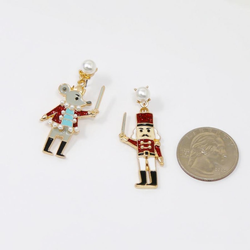 May include: A pair of festive dangle earrings featuring a mouse and a nutcracker soldier. The mouse is grey with a red jacket and a sword. The nutcracker is red, white, and black with a sword. Both have pearl accents.
