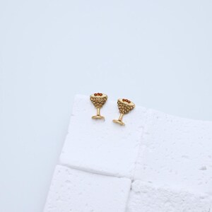 以下が含まれることがあります： 金色のマティーニ型のイヤリングのペア。各イヤリングには、小さなキラキラ光るクリスタルがちりばめられており、オリーブに似た3つの小さな赤いビーズが付いています。イヤリングは白い表面に表示されています。
