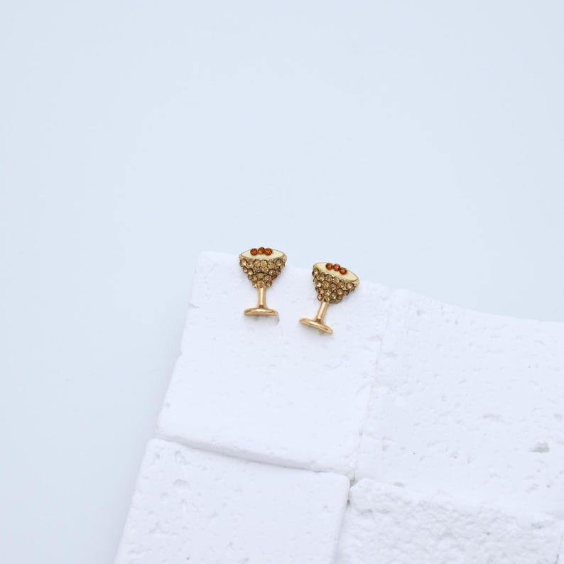 以下が含まれることがあります： 金色のマティーニ型のイヤリングのペア。各イヤリングには、小さなキラキラ光るクリスタルがちりばめられており、オリーブに似た3つの小さな赤いビーズが付いています。イヤリングは白い表面に表示されています。