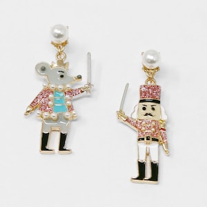 May include: A pair of novelty dangle earrings. One earring depicts a mouse in a pink jacket, holding a sword. The other earring is a nutcracker soldier in a red jacket, also holding a sword. Both earrings have pearl accents and gold-tone metal.