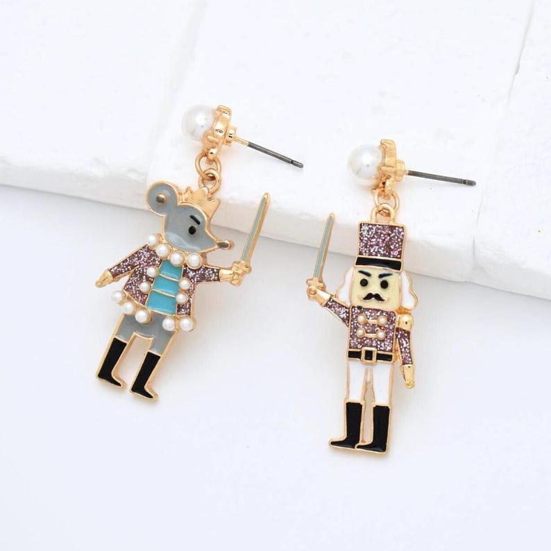 May include: A pair of gold-coloured earrings with a mouse and a nutcracker design. The mouse is grey with a gold crown and a blue shirt. The nutcracker is white with a black hat and boots. Both hold swords and are adorned with faux pearls.