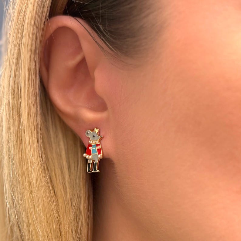 May include: A gold-toned stud earring featuring a cartoon mouse character wearing a red coat, blue vest, and a gold crown. The mouse has gray fur and black boots. The earring is a whimsical accessory.