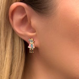 May include: Gold-tone mouse stud earrings. The earrings feature a gray mouse wearing a pink cape with white trim, a blue shirt, and black boots. The mouse has a gold crown. The earrings are set against a blurred background.